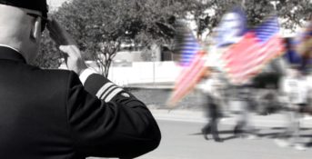 A black and white photo of a veteran saluting two American flags, which are in color