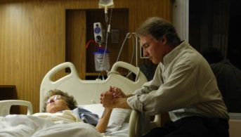 A man sits by his wife's hospital bedside, holding her hand