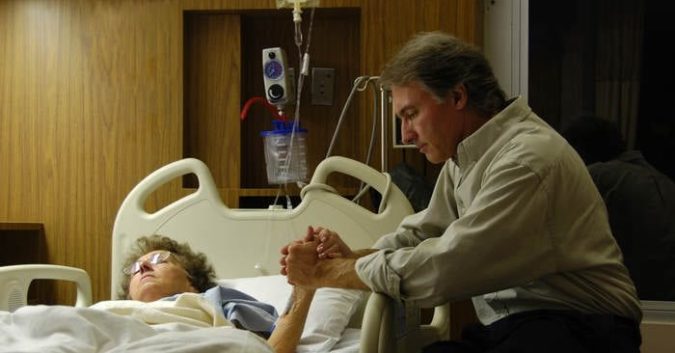 A man sits by his wife's hospital bedside, holding her hand